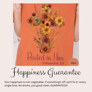 K&ouml;nnte beinhalten: Orangefarbenes T-Shirt mit floralem Kreuz-Design und dem Text "Rooted in Him" und "Colossians 2:7". Der Text "Happiness Guarantee" ist ebenfalls sichtbar.