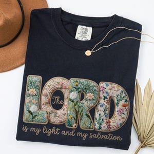 May include: Navy blue t-shirt with the embroidered word "LORD" in floral design. The text "The is my light and my salvation" is below. A gold necklace and a brown hat are also visible.