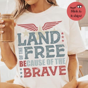 May include: A white t-shirt with a red, white, and blue graphic design. The graphic features a red, white, and blue eagle with wings spread, stars, and the text "Land of the Free Because of the Brave".