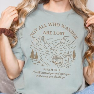 May include: A light blue t-shirt with a graphic of a vintage van driving on a winding road through a mountain landscape. The text "NOT ALL WHO WANDER ARE LOST" is printed in a brown font around the graphic. The text "WHEREVER YOU LEAD" is printed on a signpost in the graphic. The text "PSALM 32:8 I will instruct you and teach you in the way you should go." is printed below the graphic.