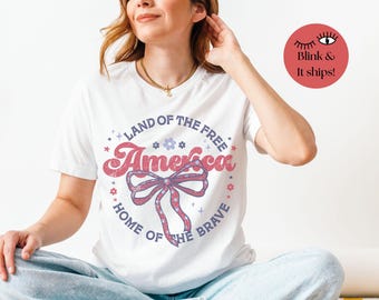 May include: White t-shirt with a patriotic design. The shirt features the word "America" in a red, retro font, surrounded by a circular design with the text "Land of the Free, Home of the Brave." A red, white, and blue bow is in the center.