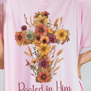 K&ouml;nnte beinhalten: Hellrosa T-Shirt mit floralem Kreuzdesign in Gelb-, Orange- und Burgundert&ouml;nen. Der Text "Rooted in Him" und "Colossians 2:7" sind darunter gedruckt. Das Wort "Blossom" befindet sich in einem rosa Kreis oben rechts.
