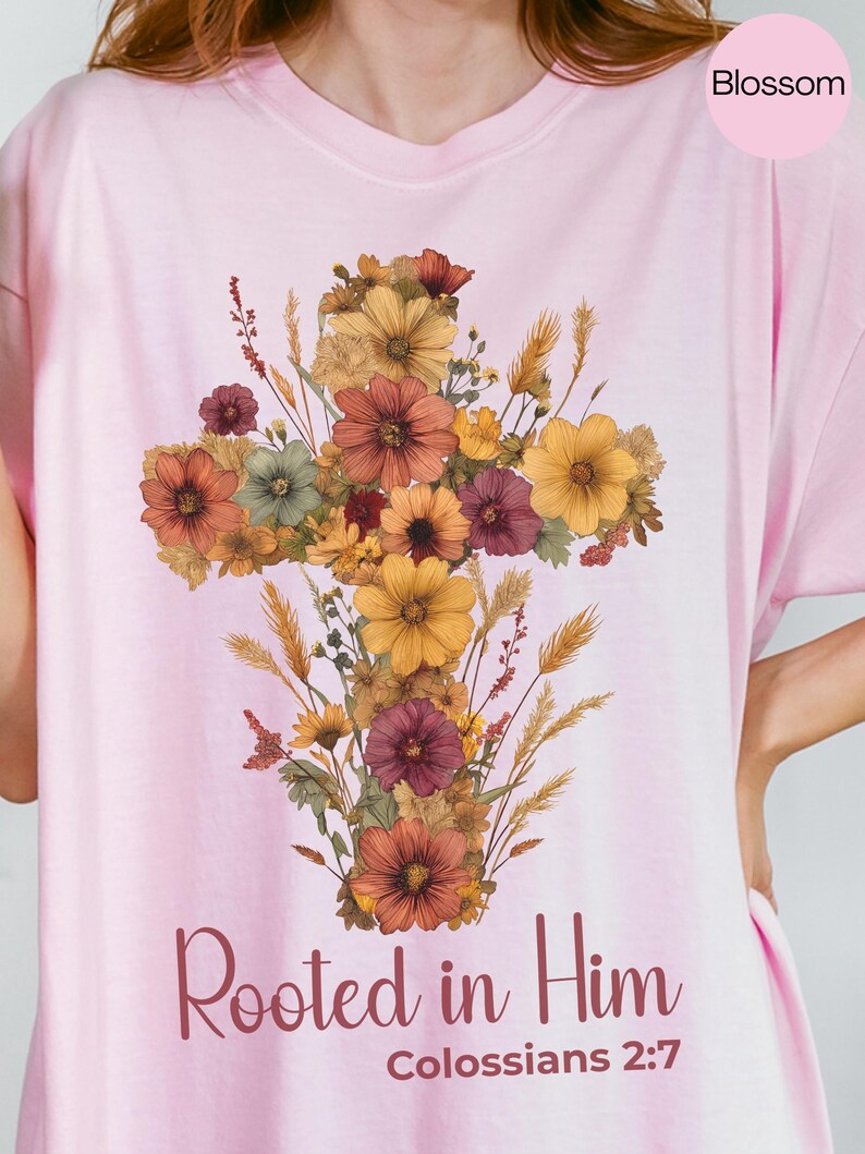 K&ouml;nnte beinhalten: Hellrosa T-Shirt mit floralem Kreuzdesign in Gelb-, Orange- und Burgundert&ouml;nen. Der Text "Rooted in Him" und "Colossians 2:7" sind darunter gedruckt. Das Wort "Blossom" befindet sich in einem rosa Kreis oben rechts.