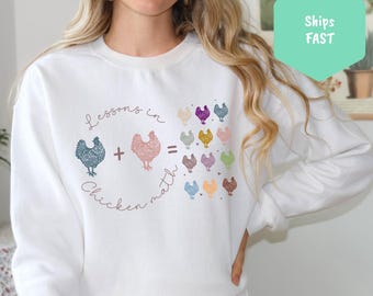 Może przedstawiać: Biała bluza z nadrukiem kurczaków i tekstem "Lessons in Chicken Math". Grafika przedstawia dwa kurczaki z plusem między nimi i szereg kurczaków w różnych kolorach.