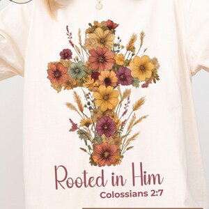 K&ouml;nnte beinhalten: Elfenbeinfarbenes T-Shirt mit einem floralen Kreuzdesign in Orange-, Gelb- und Burgundert&ouml;nen. Der Text "Rooted in Him" und "Colossians 2:7" ist darunter gedruckt. Das Shirt hat einen Rundhalsausschnitt und lange &Auml;rmel.