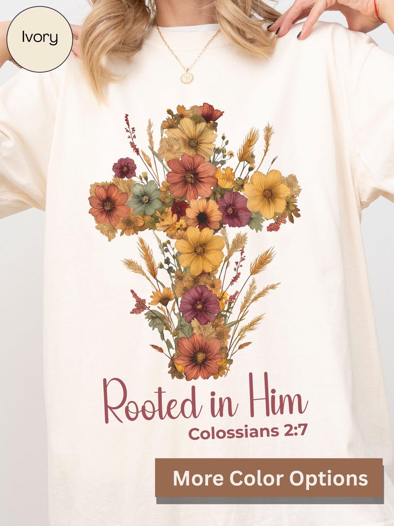 K&ouml;nnte beinhalten: Elfenbeinfarbenes T-Shirt mit einem floralen Kreuzdesign in Orange-, Gelb- und Burgundert&ouml;nen. Der Text "Rooted in Him" und "Colossians 2:7" ist darunter gedruckt. Das Shirt hat einen Rundhalsausschnitt und lange &Auml;rmel.