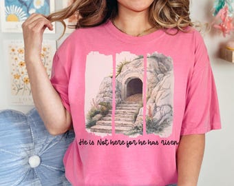May include: Pink t-shirt with a watercolor illustration of a stone tomb with an open doorway and steps leading up to it. The text "He is Not here for he has risen" is printed below the image.
