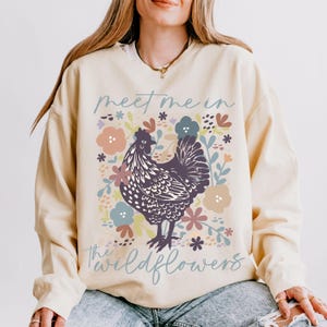 May include: A cream-colored sweatshirt with a black and white chicken illustration surrounded by colorful flowers. The text "meet me in the wildflowers" is printed on the front of the sweatshirt.
