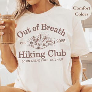 May include: White t-shirt with a brown graphic of mountains and the text "Out of Breath est 2025 Hiking Club GO ON AHEAD I WILL CATCH UP"