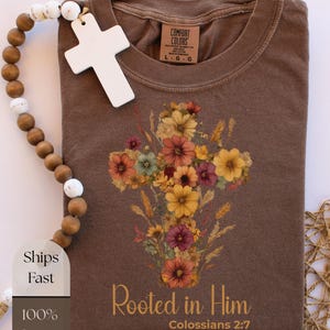 K&ouml;nnte beinhalten: Braunes T-Shirt mit floralem Kreuzdesign in Orange-, Gelb- und Lilat&ouml;nen. Der Text "Rooted in Him" und "Colossians 2:7" ist darunter gedruckt. Das Shirt besteht aus 100 % Baumwolle.