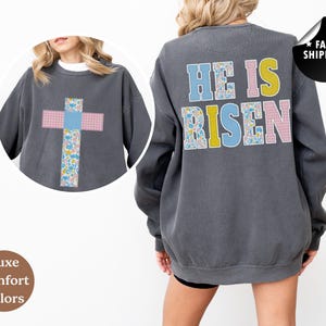 May include: Dark grey sweatshirt featuring a cross design on the front and the words "HE IS RISEN" on the back. The cross and lettering use floral and plaid patterns in pastel colours. The sweatshirt has a relaxed fit.