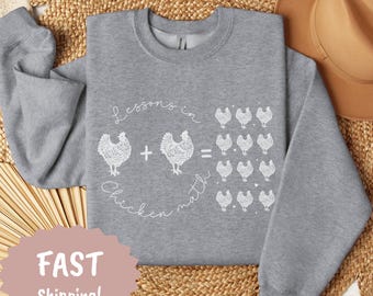 Może przedstawiać: Szara bluza z napisem "Lessons in Chicken Math" i grafiką kurczaków. Projekt przedstawia dwa kurczaki, znak plus, znak równości i grupę kurczaków. Bluza ma okrągły dekolt.