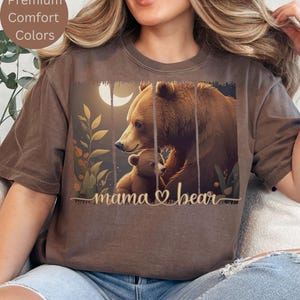 May include: A brown t-shirt with a graphic of a mama bear and her cub. The graphic is in a vintage style with a moon and leaves. The text "mama bear" is written below the graphic.