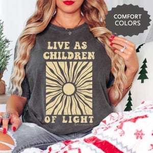 May include: Dark gray t-shirt with the text "LIVE AS CHILDREN OF LIGHT" in a beige, retro-style font. The design features a sunburst graphic. The words "Comfort Colors" are in a dark gray box in the upper right corner.