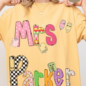 May include: Yellow Comfort Colors sweatshirt with the name "Mrs. Parker" in colorful, patterned letters. The design includes school-themed graphics like markers and a notepad. The words "More Color Options" are at the bottom.