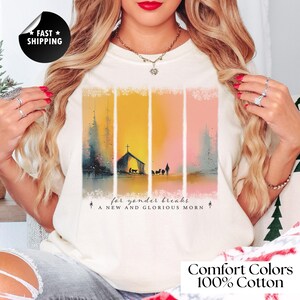 May include: Cream-colored t-shirt with a watercolor design featuring a church, people, and animals under a cross. The design is set against vertical stripes of yellow, orange, and pink. Text at the bottom reads "for yonder breaks A NEW AND GLORIOUS MORN".