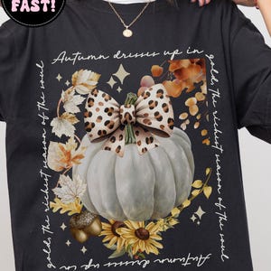 May include: Black t-shirt featuring a white pumpkin with a leopard print bow, surrounded by autumn leaves and sunflowers. The text reads "Autumn dresses up in gold, the richest season of the soul." Additional text says "More Colors Available."