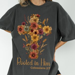 K&ouml;nnte beinhalten: Dunkelgraues T-Shirt mit einem floralen Kreuzdesign in Orange-, Gelb- und Burgundert&ouml;nen. Der Text "Rooted in Him" und "Kolosser 2:7" ist unter der floralen Anordnung in Gold gedruckt.