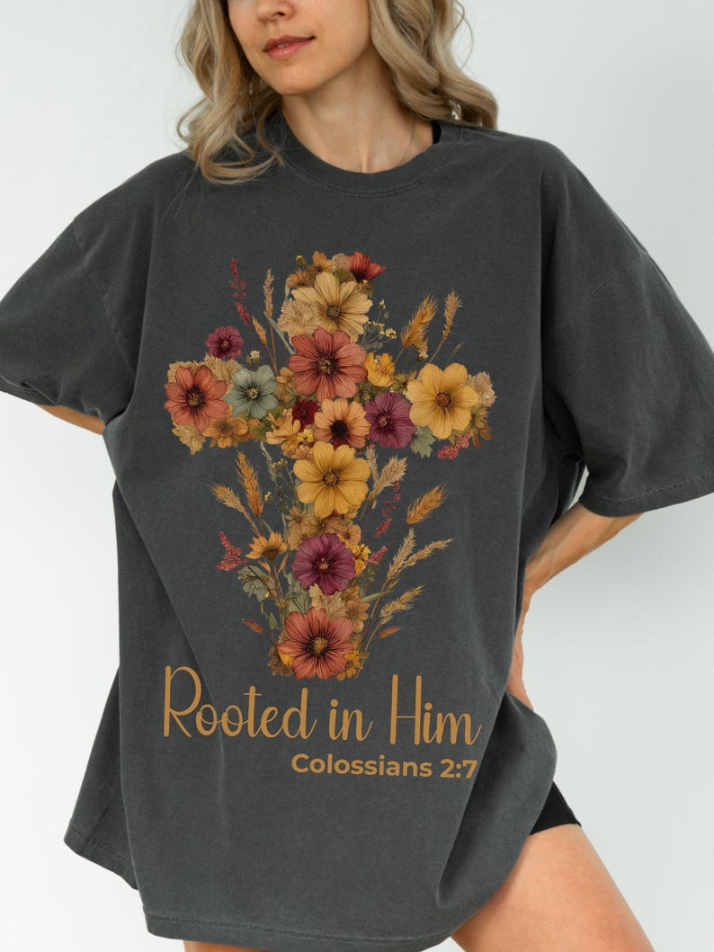 K&ouml;nnte beinhalten: Dunkelgraues T-Shirt mit einem floralen Kreuzdesign in Orange-, Gelb- und Burgundert&ouml;nen. Der Text "Rooted in Him" und "Kolosser 2:7" ist unter der floralen Anordnung in Gold gedruckt.