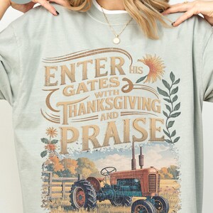 Puede incluir: Sudadera verde claro con el texto "ENTER HIS GATES WITH THANKSGIVING AND PRAISE" sobre una ilustración de un tractor vintage. El tractor es rojo y azul, ambientado en un campo con flores y una valla.