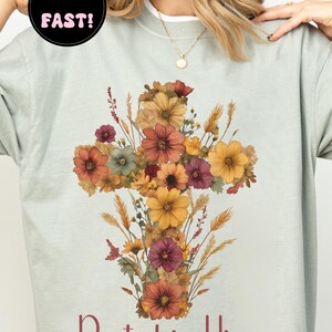 K&ouml;nnte beinhalten: Hellgr&uuml;nes Sweatshirt mit einem floralen Kreuzdesign in Orange, Gelb und Burgunderrot. Der Text "Rooted in Him" und "Kolosser 2:7" sind darunter aufgedruckt. Das Sweatshirt hat einen Rundhalsausschnitt.