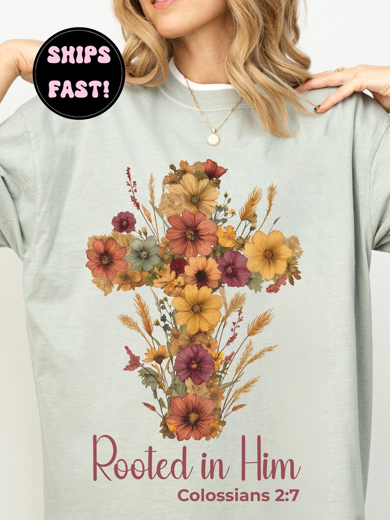 K&ouml;nnte beinhalten: Hellgr&uuml;nes Sweatshirt mit einem floralen Kreuzdesign in Orange, Gelb und Burgunderrot. Der Text "Rooted in Him" und "Kolosser 2:7" sind darunter aufgedruckt. Das Sweatshirt hat einen Rundhalsausschnitt.