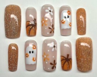 May include: Set of ten false nails with a Halloween and fall theme. The nails feature a variety of designs, including ghosts, pumpkins, and flowers. Some nails are a solid brown glitter color.