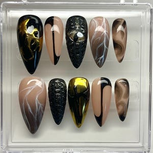 May include: A set of ten artificial nails in a clear plastic case. The nails feature various designs, including black and gold swirls, a black and tan split design, and a textured black nail with gold accents. Other nails have tan and white marbled designs.