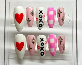 May include: A set of ten false nails in a clear plastic case. The nails are decorated with hearts, XOXO, and pink and white gingham patterns.
