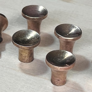 May include: A collection of six antique brass cabinet knobs. One knob has a screw attached. The knobs have a rounded top and a cylindrical base. The brass has a warm, aged patina, suggesting vintage hardware.