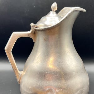 May include: A vintage silver-colored pitcher with a lid and a handle. The pitcher has a rounded body, a spout, and a small finial on the lid. The handle is angular. The pitcher sits on a small, circular base. The metal has a slightly tarnished appearance.