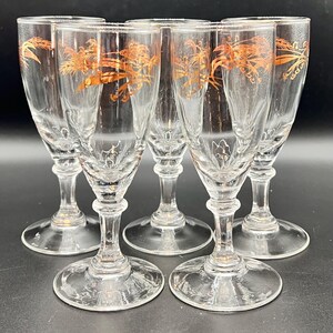 May include: Set of six clear glass cordial glasses with gold palm tree designs. Each glass has a round base, a slender stem, and a flared rim. The gold design is repeated on each glass.