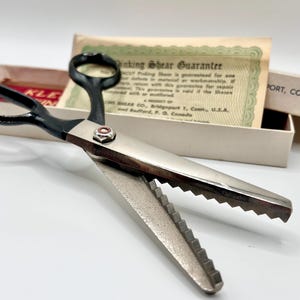 May include: Close-up of vintage pinking shears with black handles and silver blades. The blades have a zig-zag pattern. The shears are open, revealing the teeth. A guarantee card is in the background.