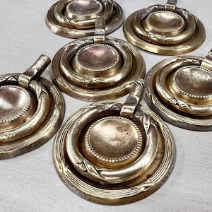 May include: A collection of antique brass drawer pulls. Each pull features a circular design with concentric rings and a small handle. The brass has a tarnished, aged appearance, suggesting vintage craftsmanship.