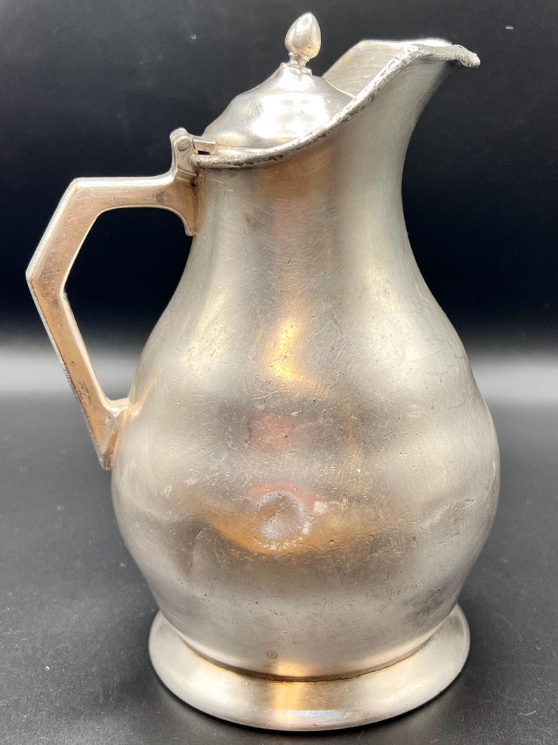 May include: A vintage silver-colored pitcher with a lid and a handle. The pitcher has a rounded body, a spout, and a small finial on the lid. The handle is angular. The pitcher sits on a small, circular base. The metal has a slightly tarnished appearance.