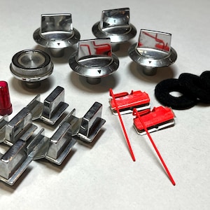 May include: A collection of vintage car parts, including chrome knobs with directional indicators, a red plastic tube, red and white rectangular pieces, and black felt washers. The parts are arranged on a white surface.