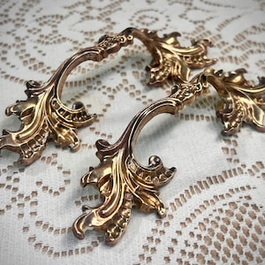 May include: Four ornate, gold-colored cabinet pulls with a leaf-like design. The pulls have a curved handle and decorative, detailed embellishments. The hardware is likely vintage or antique, adding a touch of elegance to furniture.