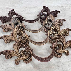 May include: Three ornate bronze drawer pulls with a scrolling leaf design. The pulls have a curved handle and are in varying shades of bronze, from light gold to dark brown. The hardware is likely for a cabinet or dresser.
