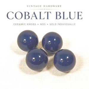 May include: Four round, glossy cobalt blue ceramic knobs arranged in a cluster. The text above reads VINTAGE HARDWARE COBALT BLUE CERAMIC KNOBS • NOS • SOLD INDIVIDUALLY. Each knob has a smooth, reflective surface with a uniform dark blue finish.