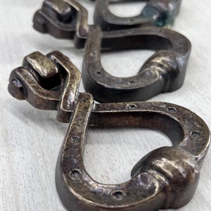 May include: Close-up of several antique bronze-colored metal belt buckles. Each buckle features a unique design with a fist-shaped end and a horseshoe-shaped loop. The buckles are arranged in a row, showcasing their intricate details and aged patina.