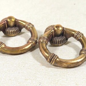 May include: Two circular brass drawer pulls featuring a hinged ring design. Each pull has a central mounting point with a ribbed, fan-like base and a smooth, rounded ring that includes decorative horizontal bands along its circumference.