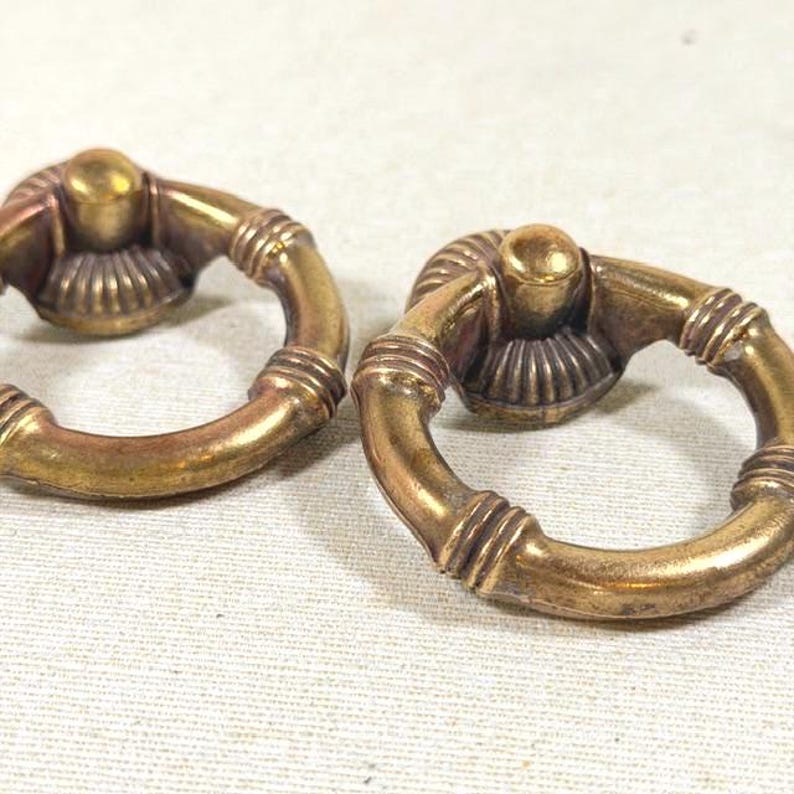 May include: Two circular brass drawer pulls featuring a hinged ring design. Each pull has a central mounting point with a ribbed, fan-like base and a smooth, rounded ring that includes decorative horizontal bands along its circumference.