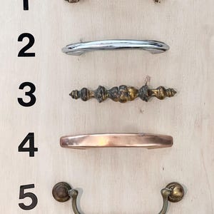 May include: Five different cabinet pulls are displayed against a light wood background. The pulls vary in style and finish, including a textured brass pull, a chrome pull, an antique brass pull, a copper pull, and a brass pull with a curved handle.