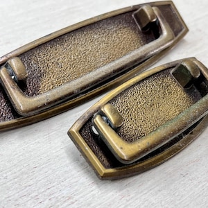 May include: Two antique brass drawer pulls with a rectangular backplate and a curved handle. The pulls have a textured surface and a warm, aged patina, suggesting vintage or retro style. The hardware is likely for furniture restoration.