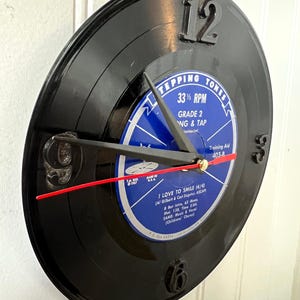 May include: A black vinyl record clock with a blue and white label. The clock features black numbers at 12, 3, 6, and 9 o'clock, with black clock hands and a red second hand. The label reads "STEPPING TONES" and "I LOVE TO SMILE".
