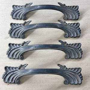 May include: Four antique metal drawer pulls with a dark gray finish. Each pull features a curved handle with ornate, stylized leaf and scroll detailing. The pulls are arranged in a vertical stack, showcasing their decorative design.