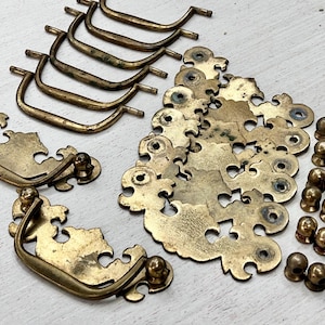 May include: A collection of antique brass hardware components, including drawer pulls, backplates, and knobs. The drawer pulls are curved with a decorative design, while the backplates have an ornate, cut-out pattern. The knobs are small and rounded. The hardware has a vintage aesthetic.