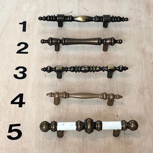 May include: Five antique-style cabinet pulls in various designs. The pulls are made of metal with a bronze finish. One pull has a white center. The pulls are arranged vertically on a light wood surface.