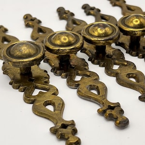May include: A collection of antique brass drawer pulls. Each pull features a round knob and an ornate, decorative backplate. The brass has a warm, aged patina, suggesting vintage or vintage-inspired hardware. The pulls are arranged in a row, showcasing their design.