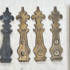 May include: Four antique brass-toned metal hinges with ornate fleur-de-lis tops and decorative floral accents. One hinge is darker, with a measurement of approximately 4 inches in length. The hinges are arranged vertically, next to a yellow measuring tape.